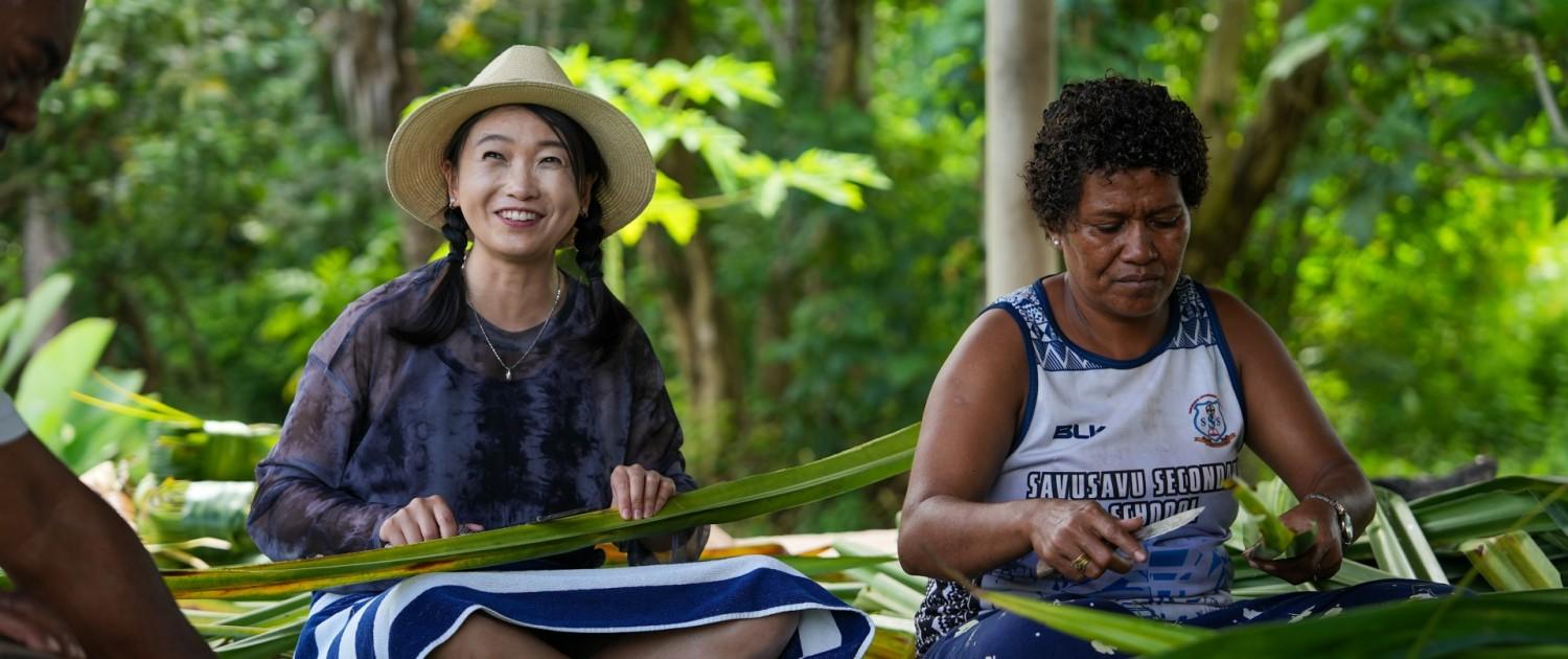A guest and a Fijian woman are sitting down and cutting pandanus leaves.